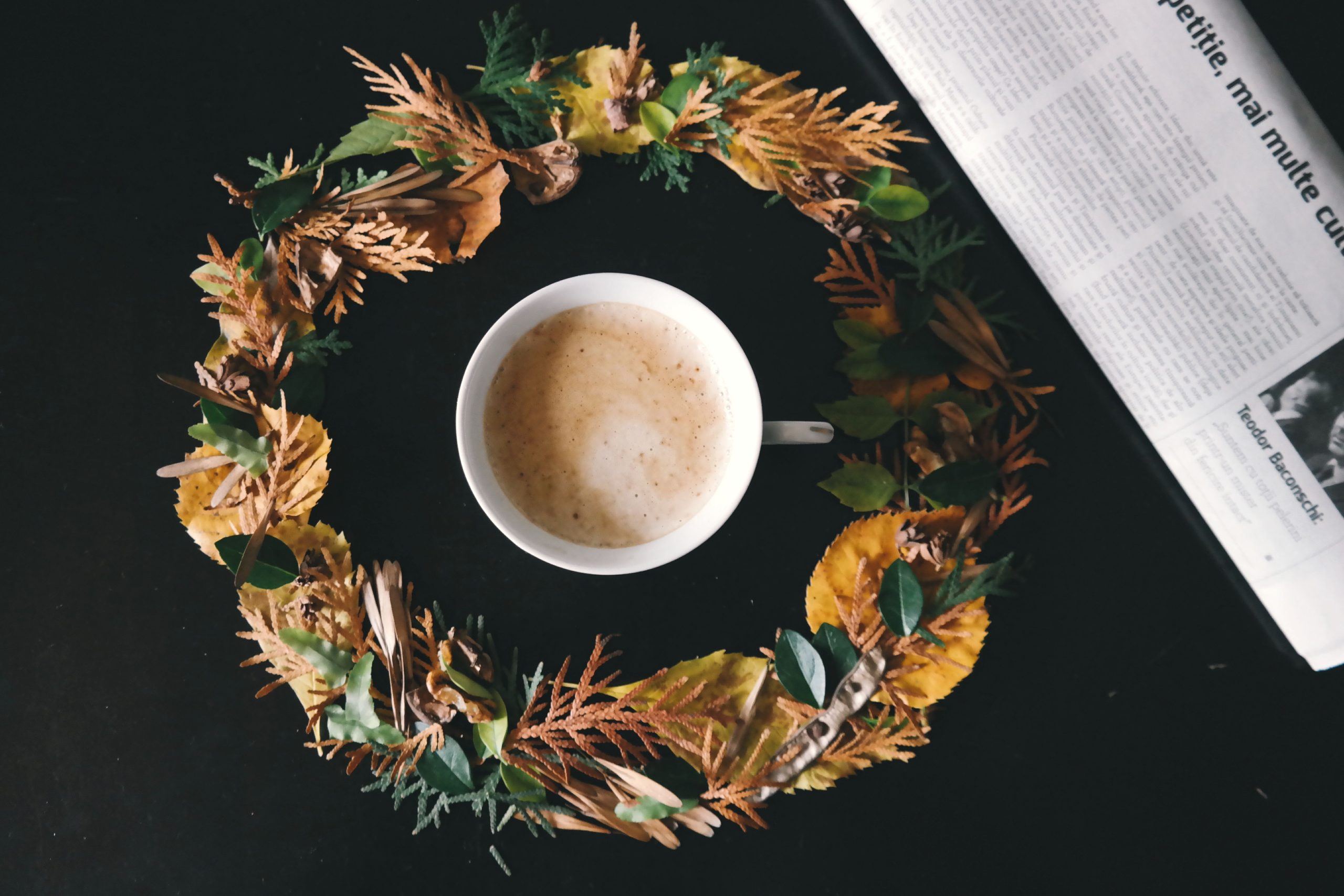 A warm cup of coffee surrounded by an autumn wreath beside a newspaper on a dark surface.