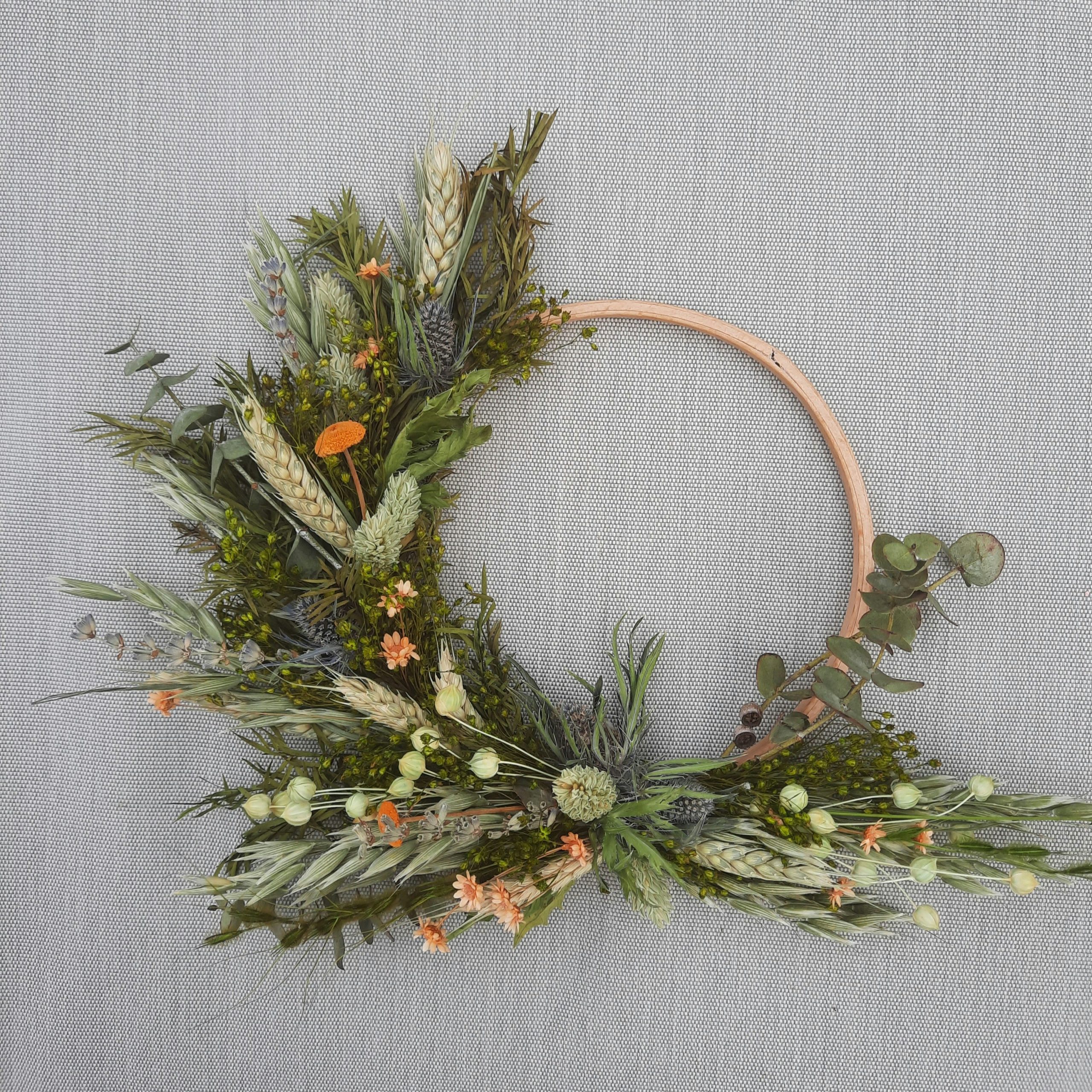 An asymmetrical dried flower wreath arranged on a natural wooden hoop, featuring wheat, eucalyptus, wild grasses, and delicate botanical accents. The lush greenery and soft earthy tones create a moder