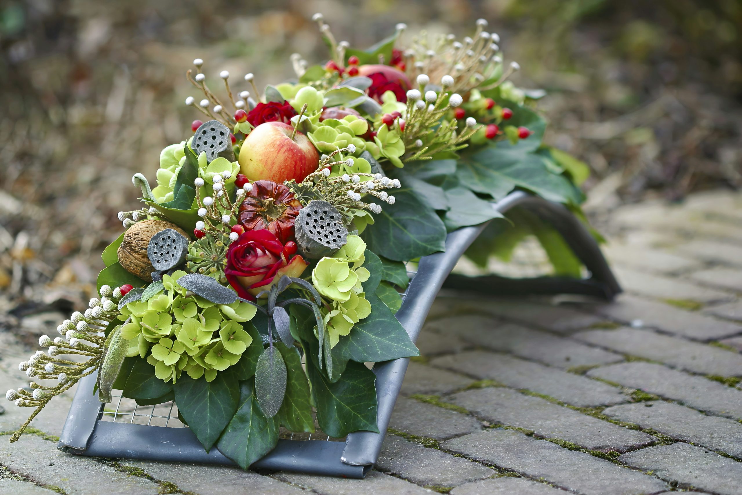 Autumn floral arrangement featuring an apple, red roses, green hydrangea, berries, and natural elements, set on a paved surface. Perfect for fall themes, remembrance, or rustic decor.