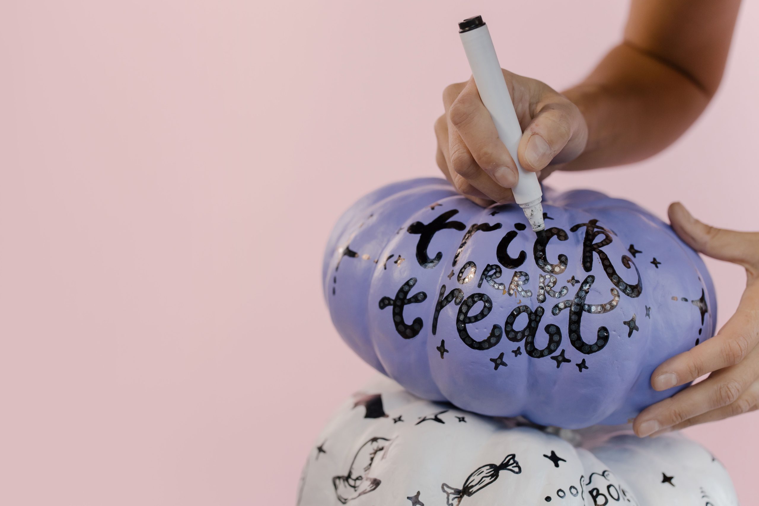 Close-up of hands decorating a blue Halloween pumpkin with 'trick or treat' on a pink background.
