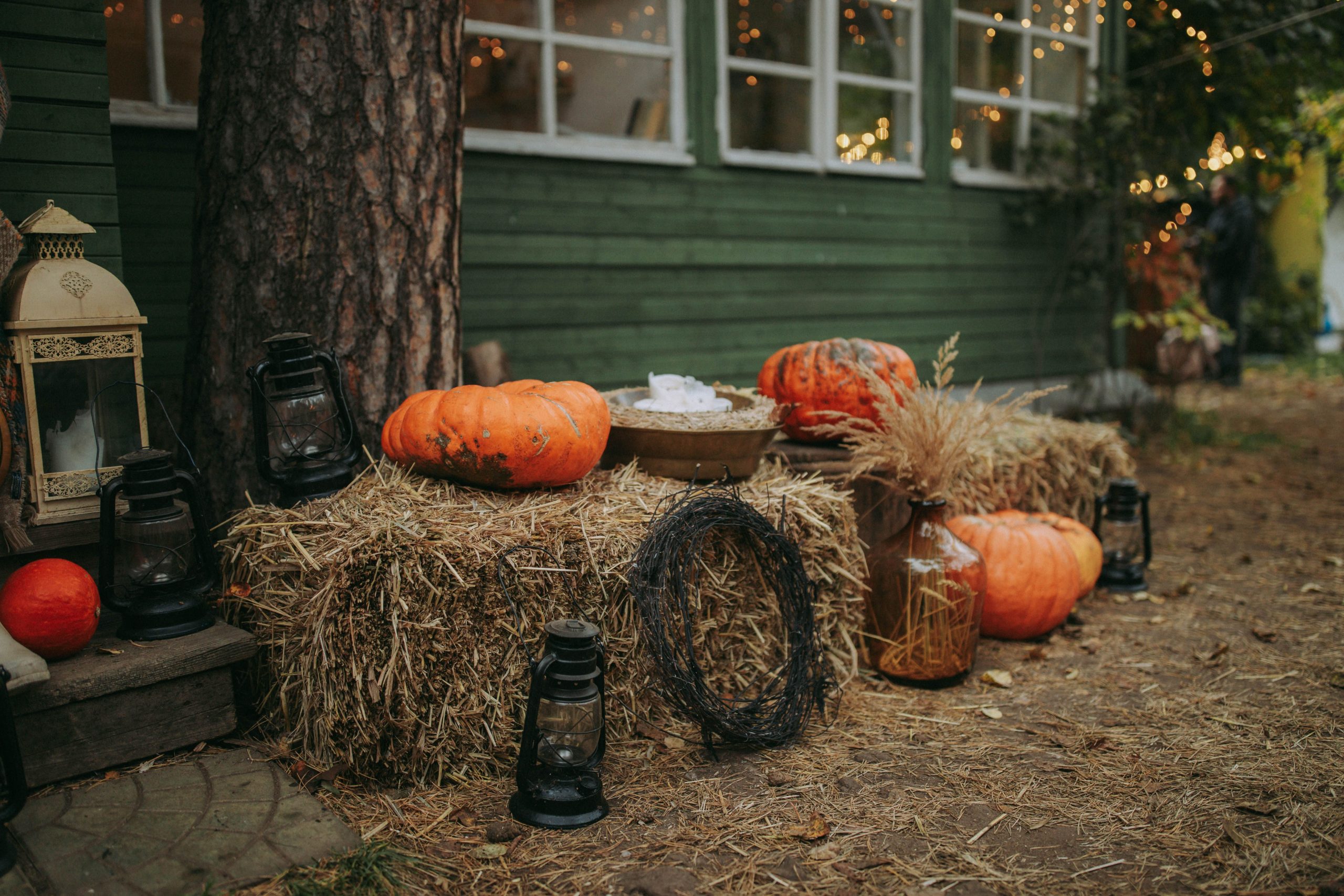 Cozy fall setting with pumpkins, hay bales, and lanterns by a wooden house.