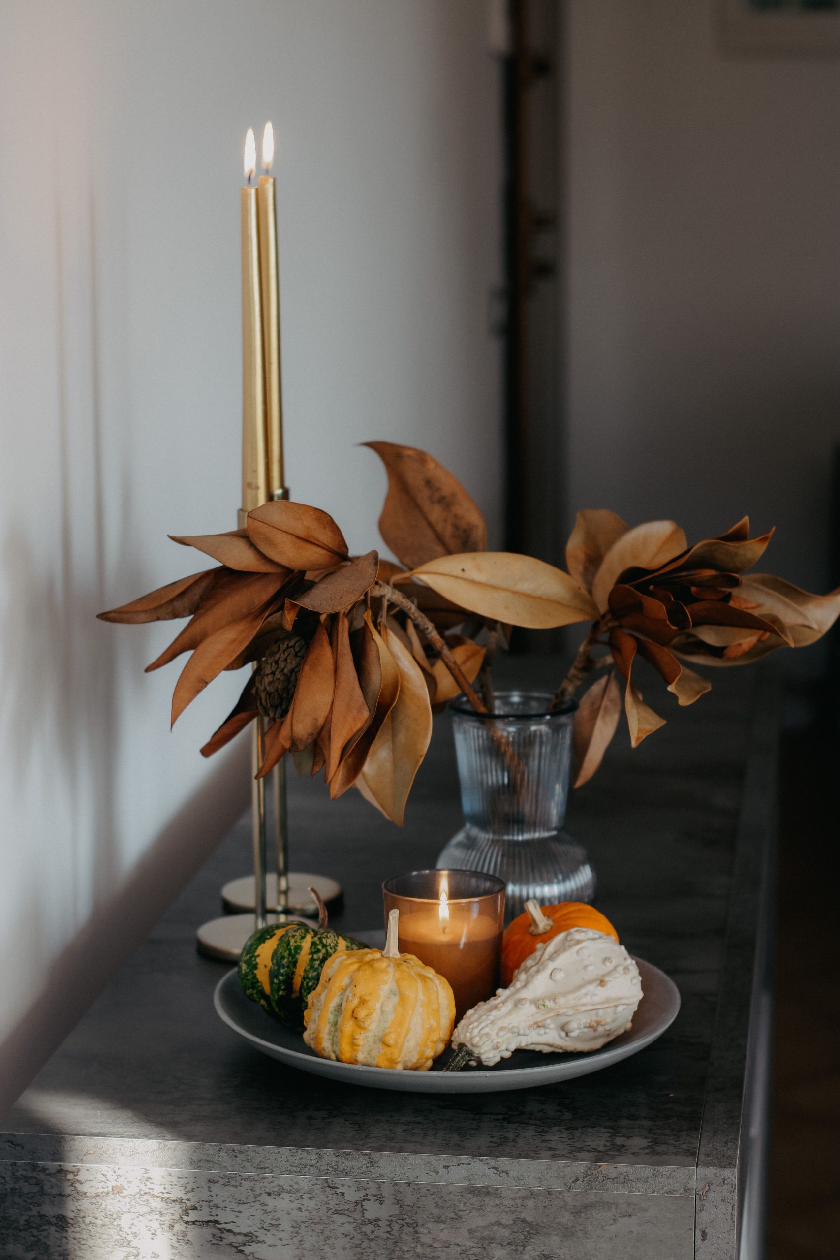 Cozy fall still life scene with candles, dried leaves, and decorative gourds on a minimalist table.
