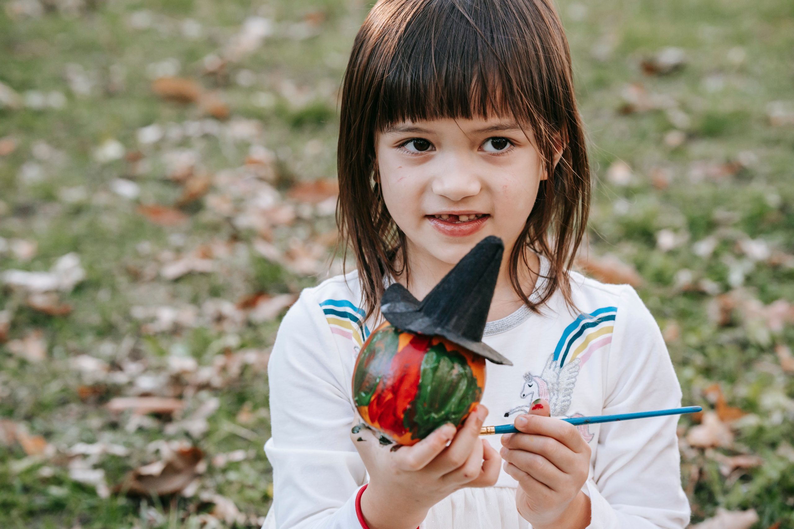 Crop cute little girl with dirty fingers painting pumpkin in witch hat for Halloween