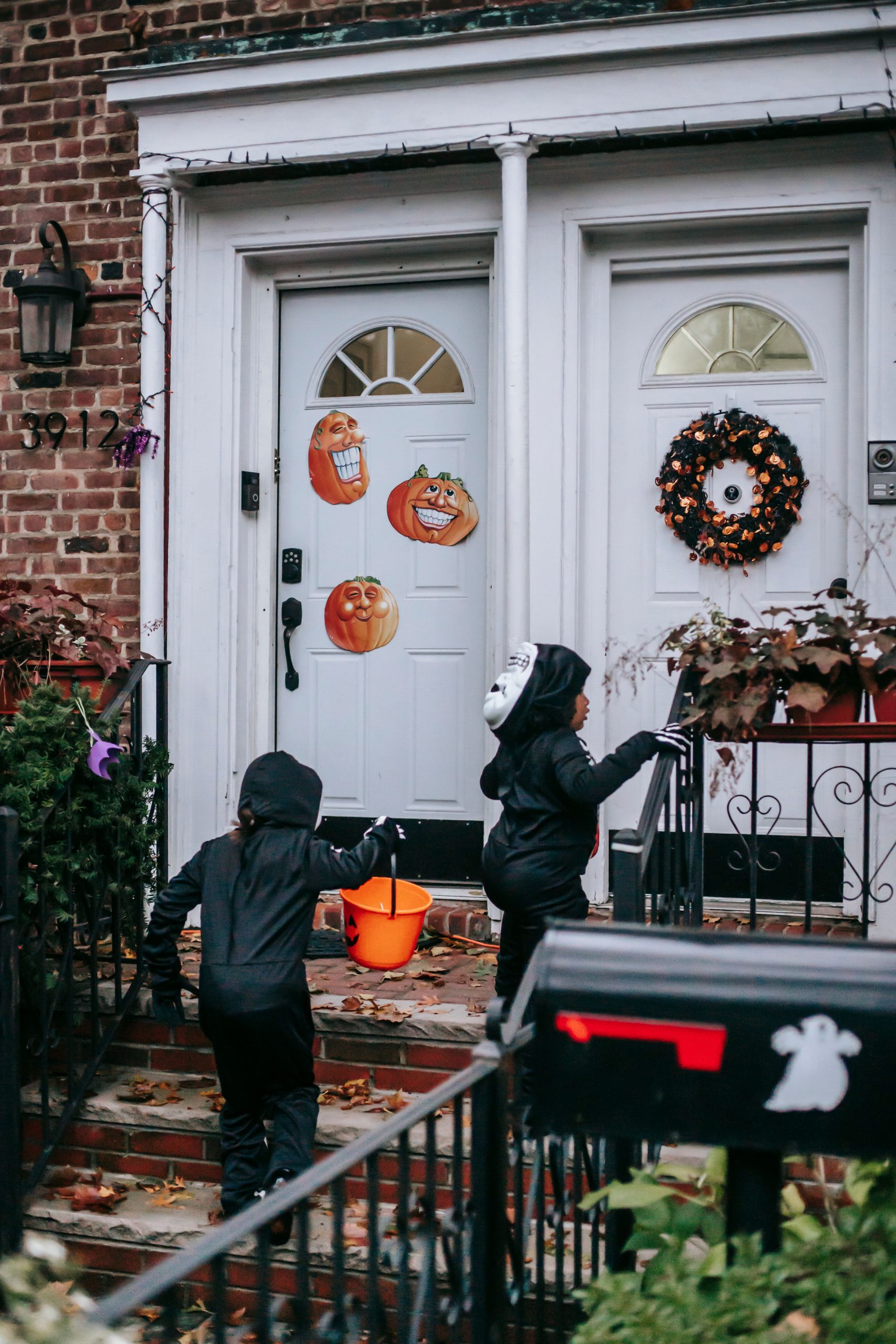 Full length of anonymous kids in black Halloween costumes with masks walking with orange buckets on stairs near house entrance