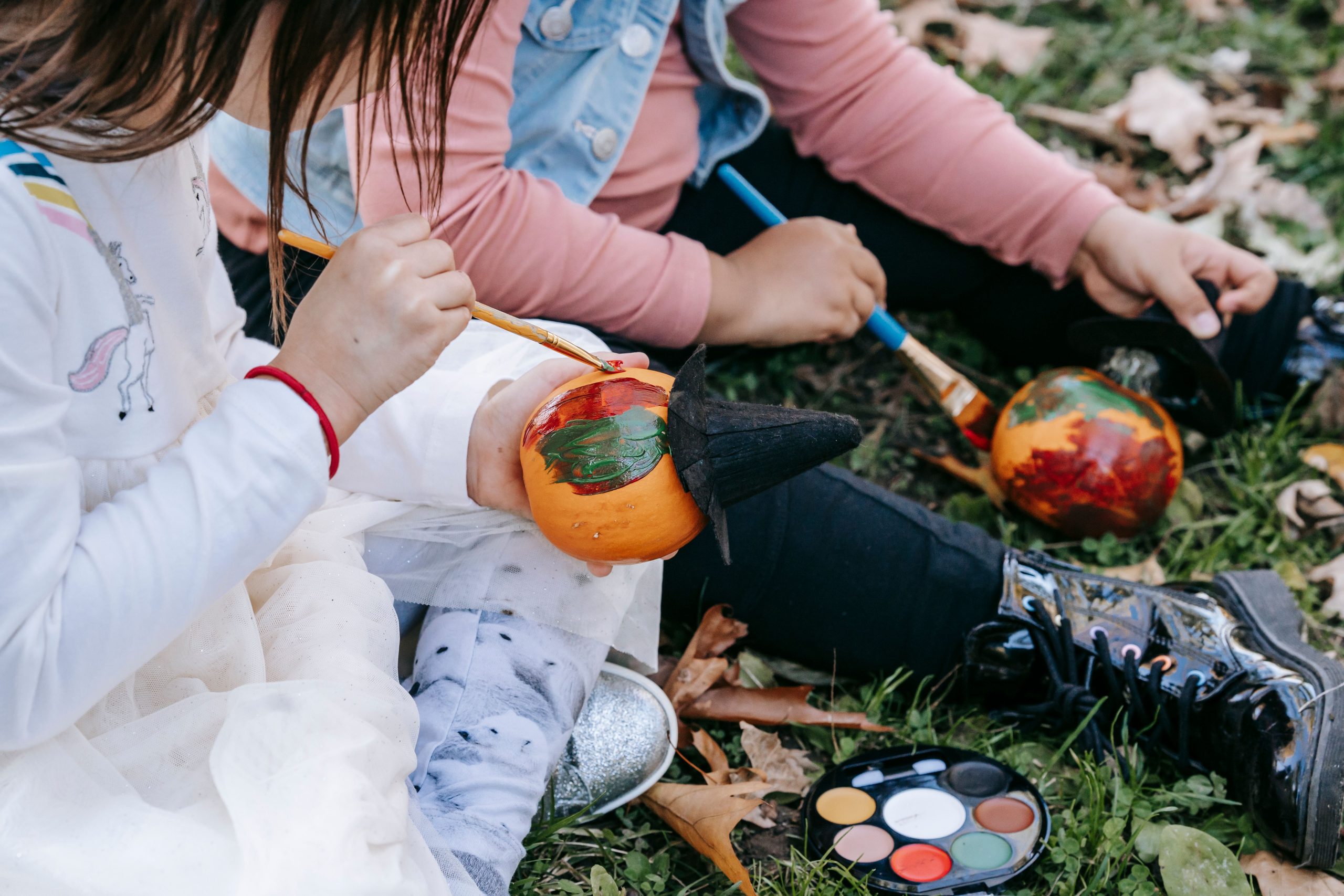 High angle of crop anonymous multiethnic girls sitting on grass and painting pumpkin with aquarelle for Halloween celebration