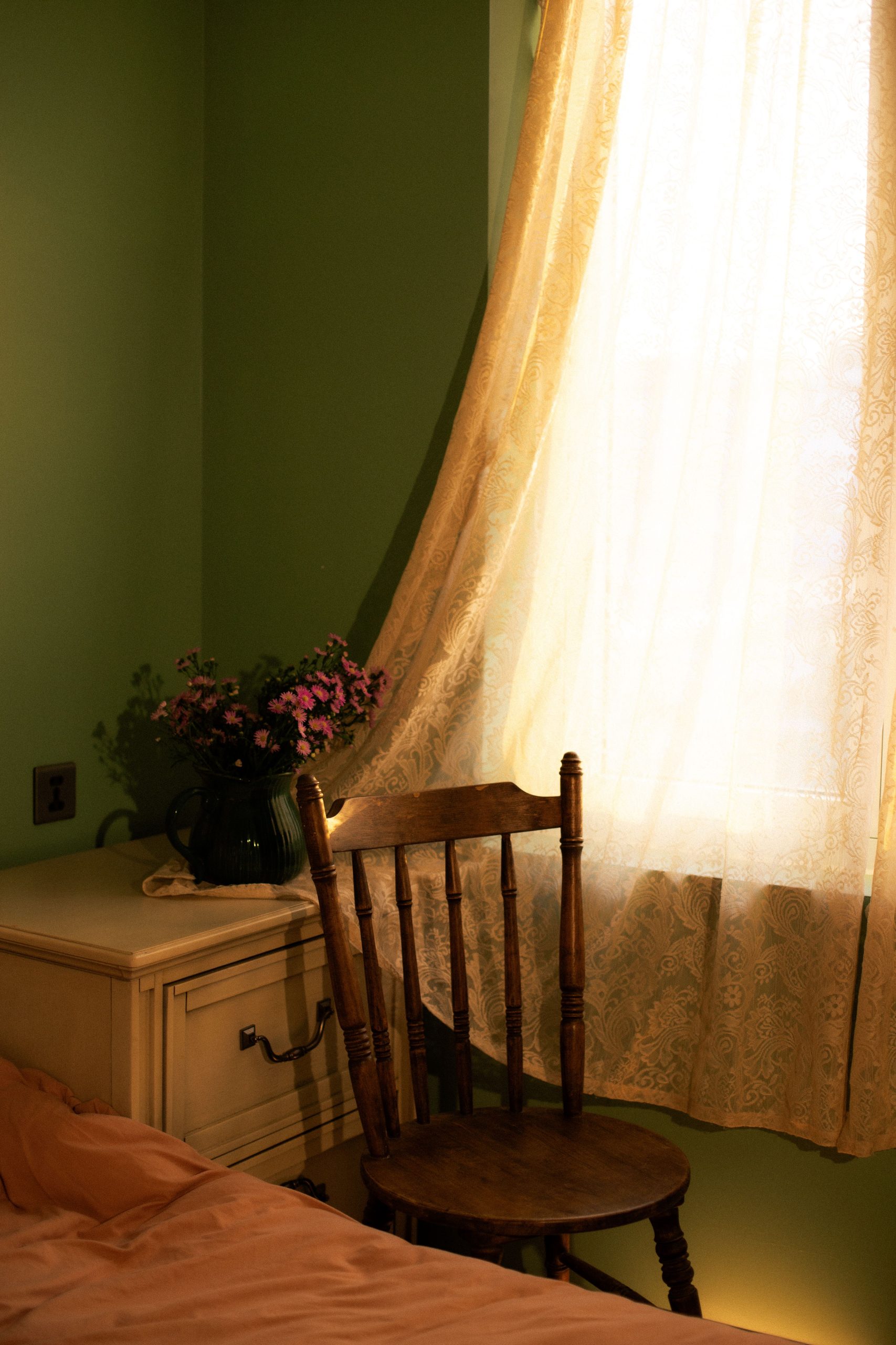 Warm and cozy bedroom corner with wooden chair, floral arrangement, and lace curtains.