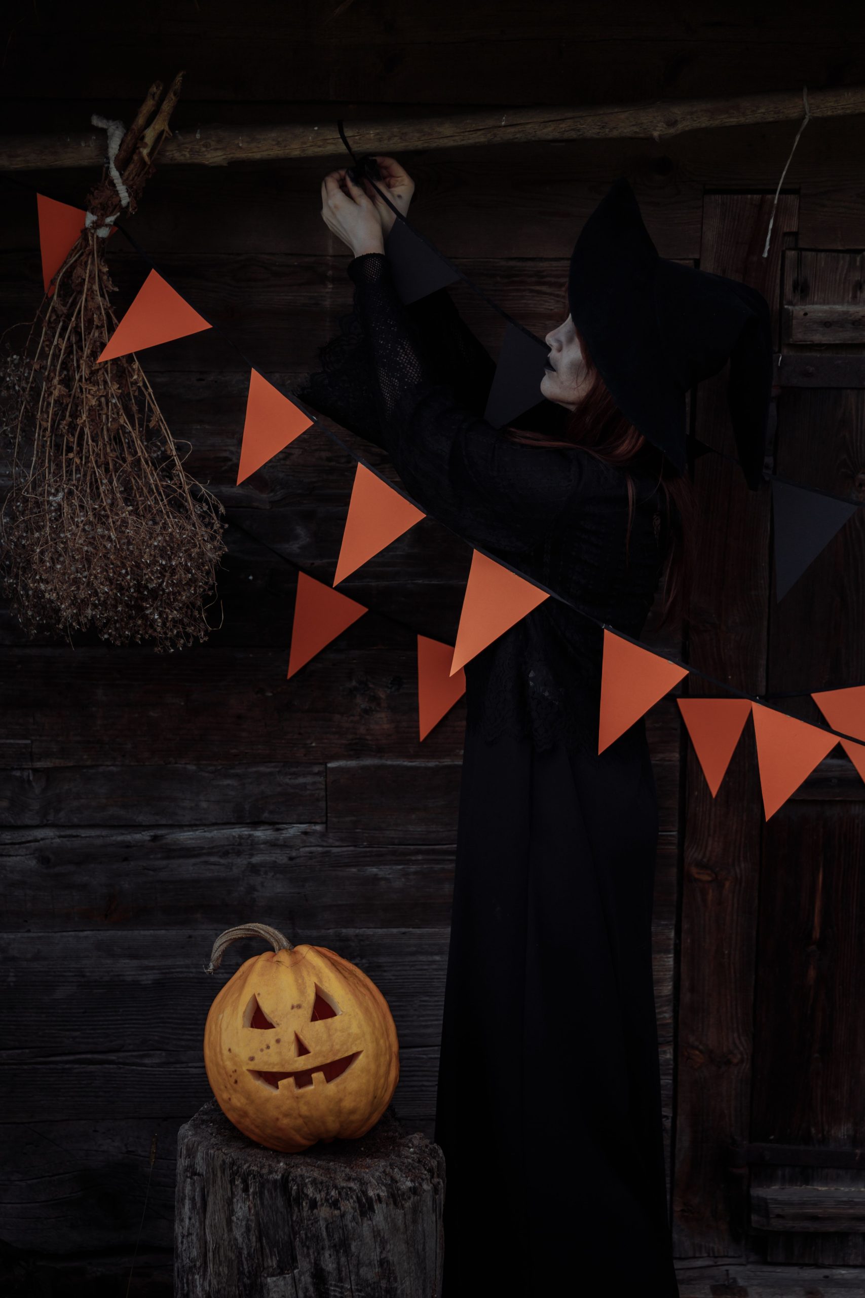 Witch in dark costume decorates woodshed with pumpkins and garlands for Halloween.