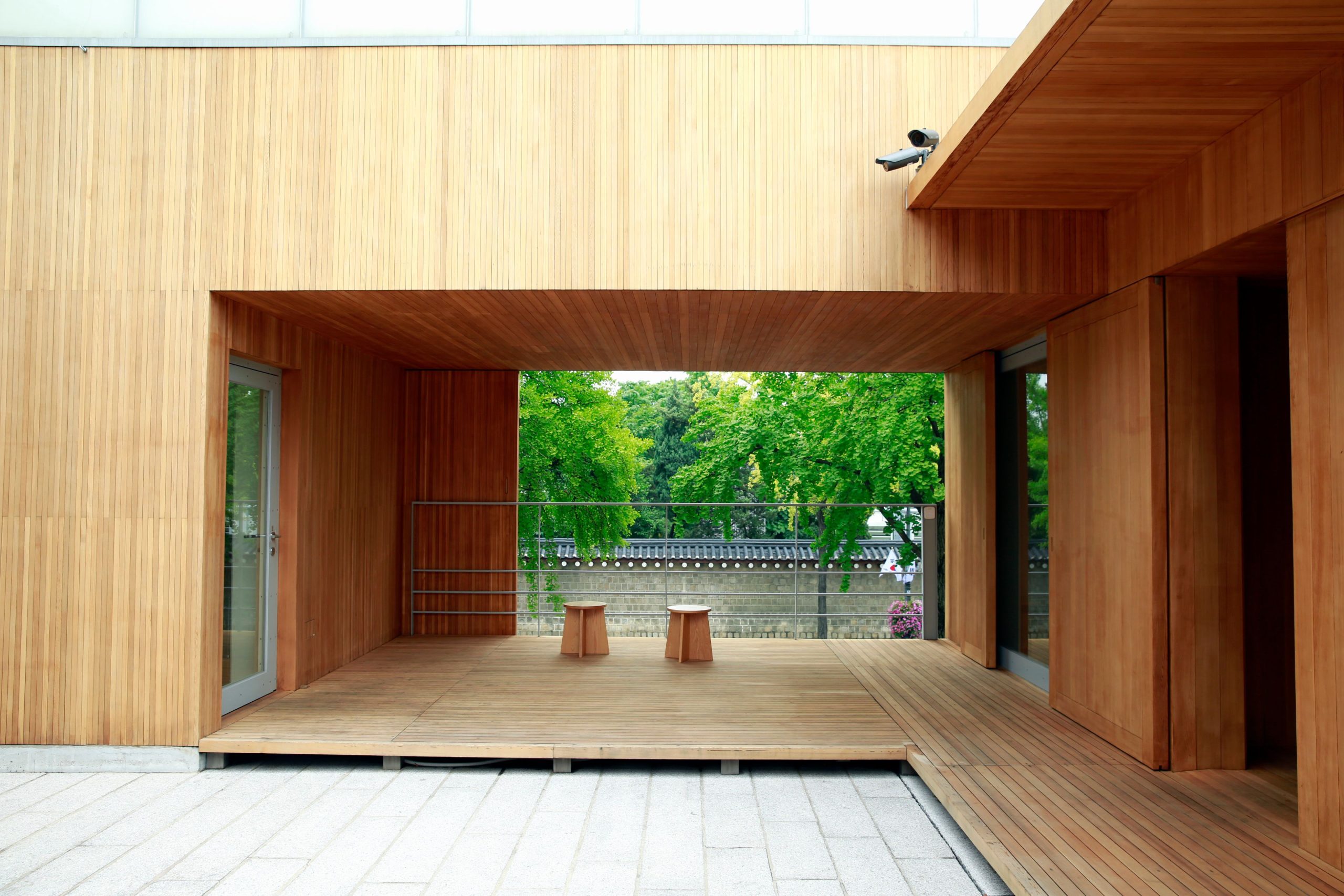 A minimalist wooden patio design with two stools and lush greenery in the background.