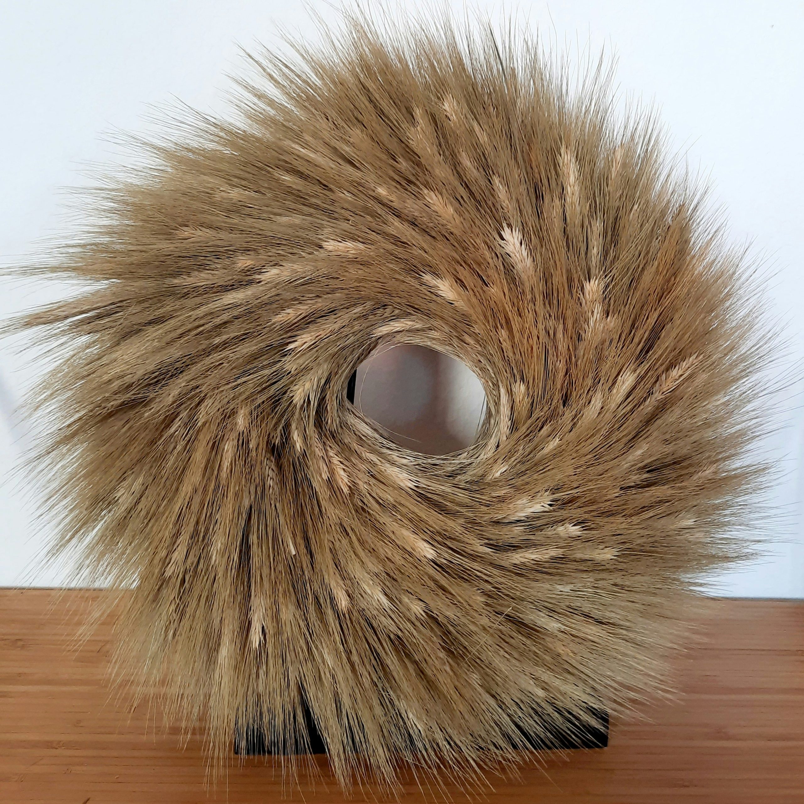 A sculptural wreath crafted from dried wheat sheaves forms a striking circular silhouette with a hollow center. The dense, radiating grain heads create rich texture and warm golden tones, set against 