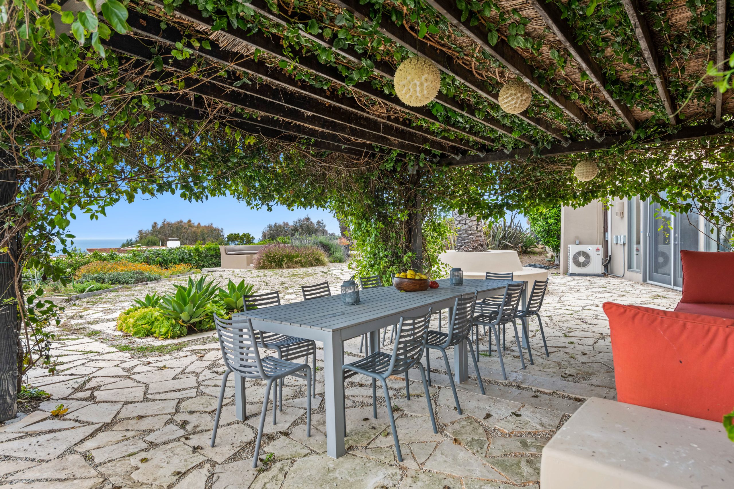 A serene garden patio with a dining setup under a vine-covered pergola, offering an ocean view.