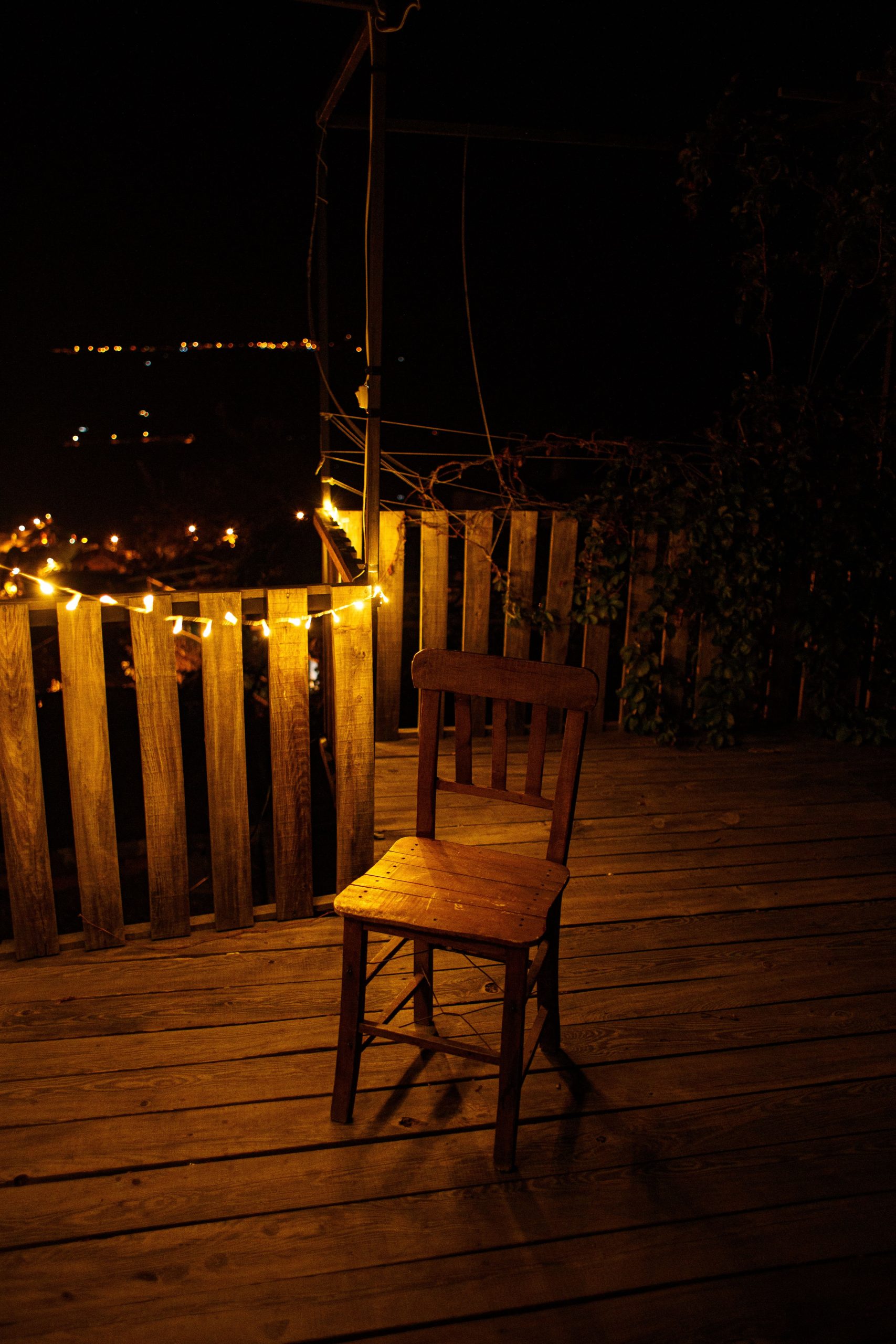 A solitary wooden chair on a deck illuminated by warm string lights at night.