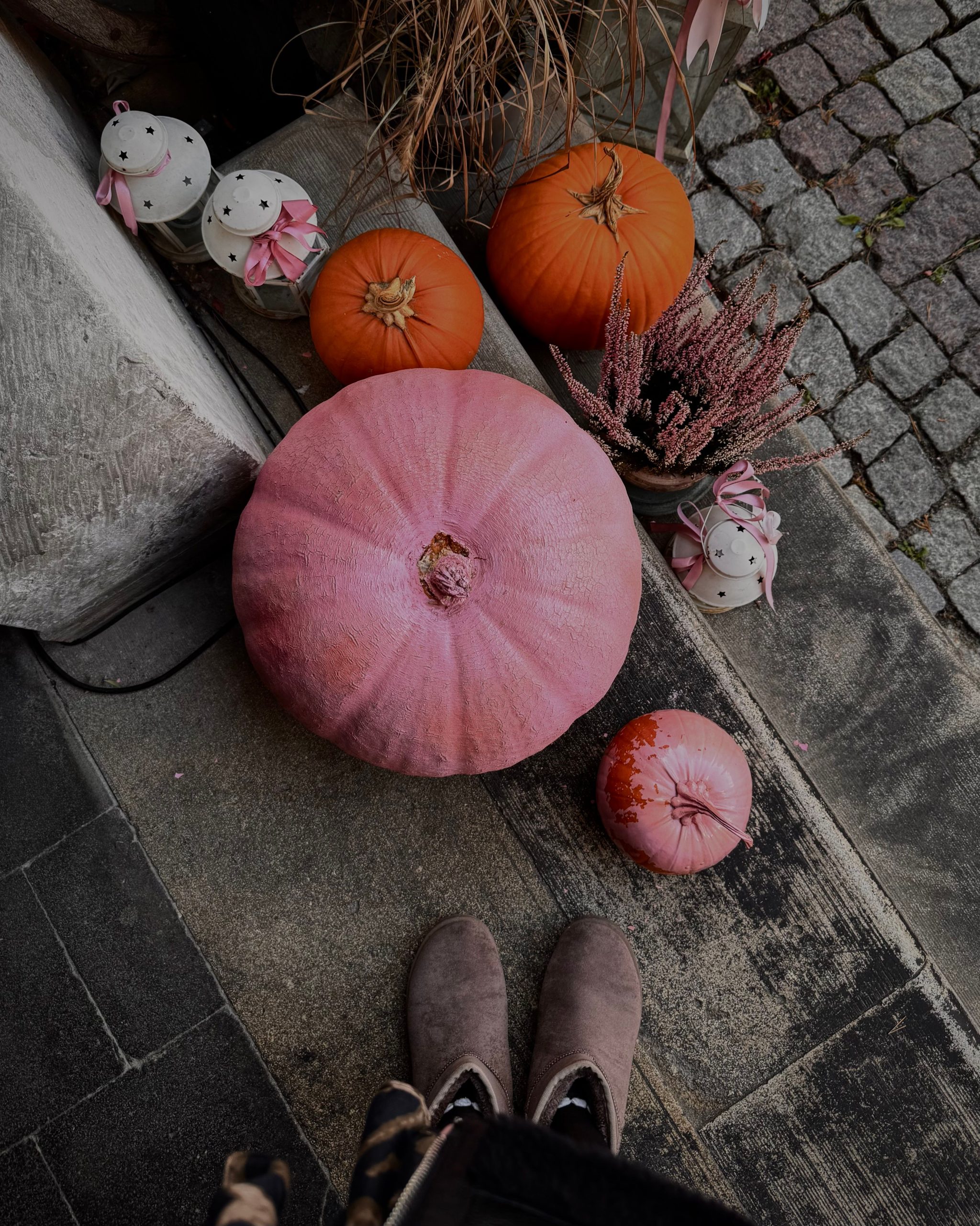 A vibrant autumn scene with pink and orange pumpkins next to decorative candles and boots on a cobblestone path.