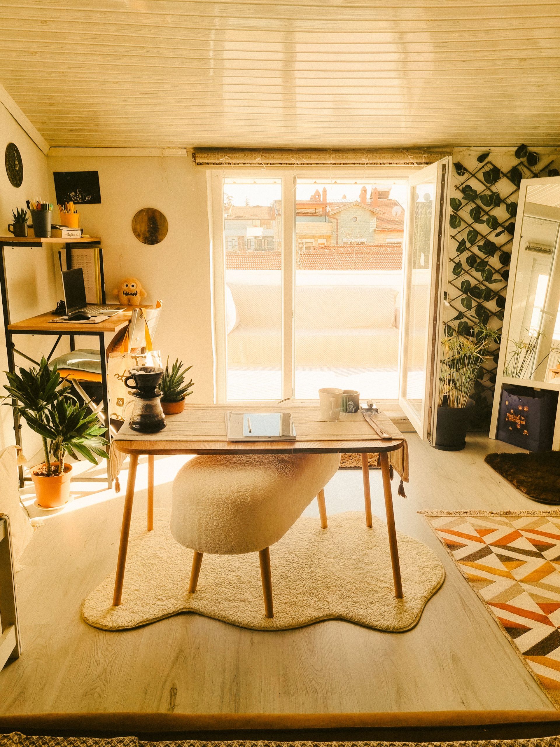 A warm, stylish home office setup featuring a desk, plants, and sunlight streaming in through large windows.