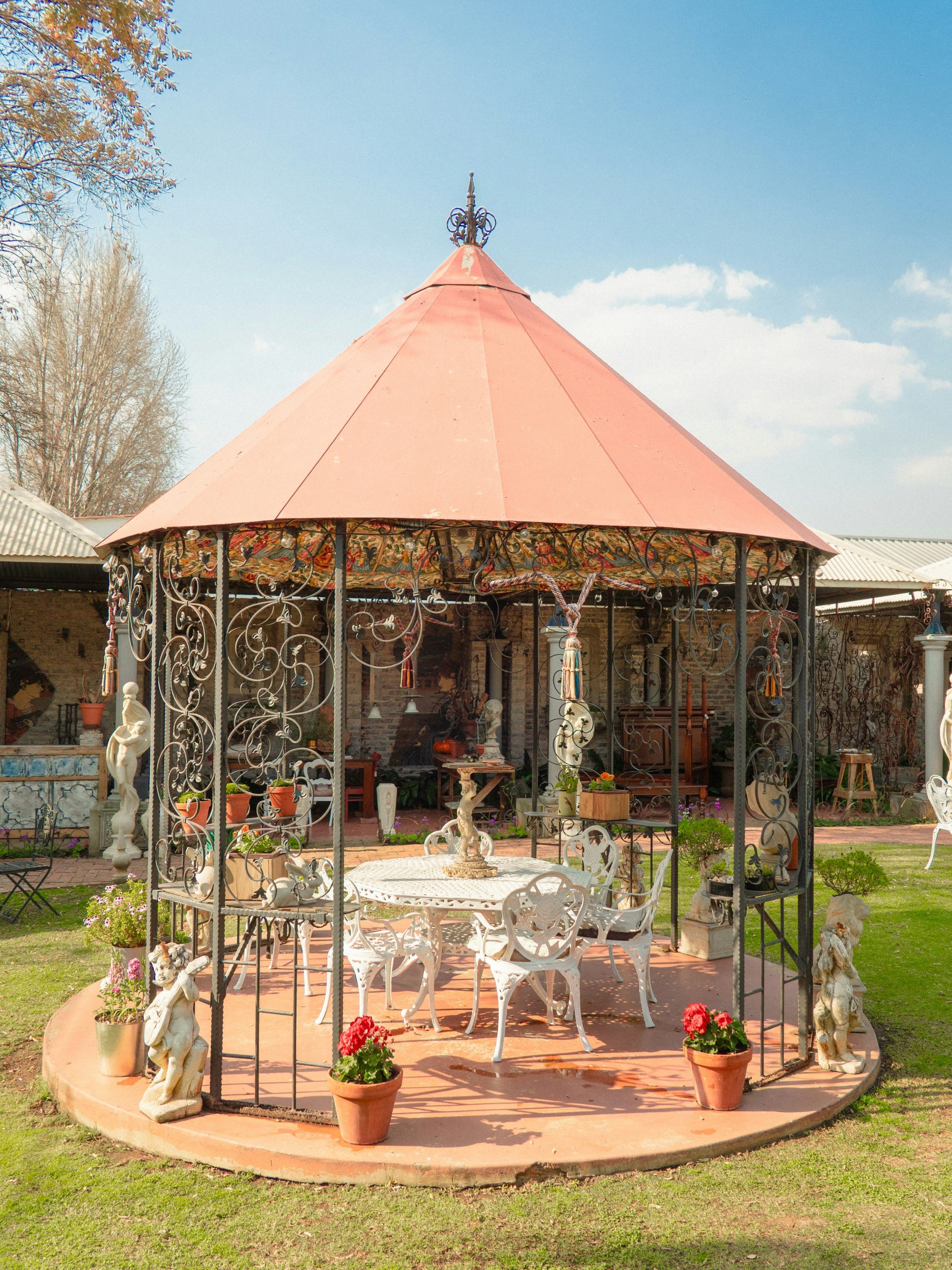 An ornate wrought-iron gazebo with a warm pink roof, white garden furniture, flowers and classical sculptures in a sunlit garden.
