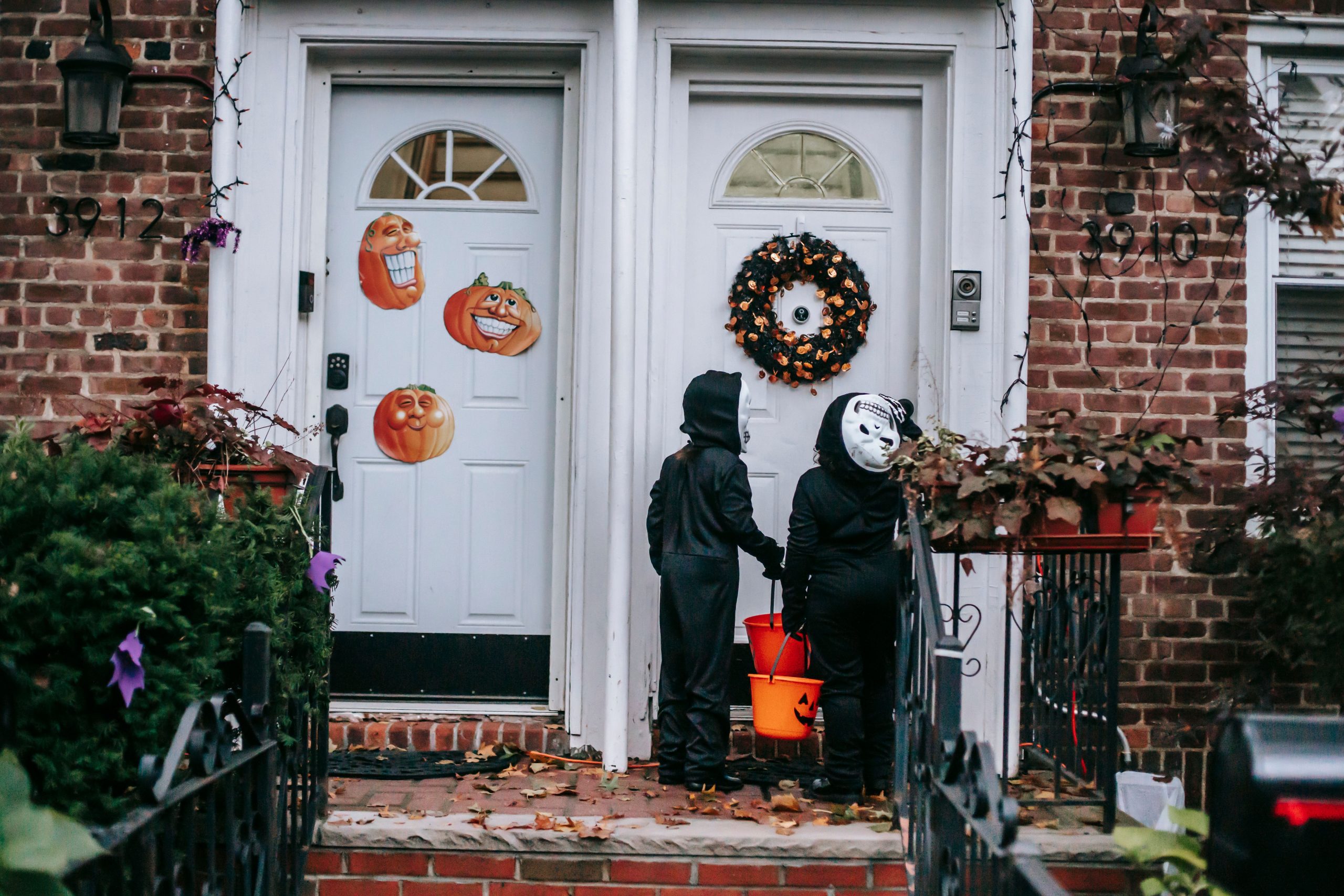 Back view full body of unrecognizable kids wearing skeleton costumes with orange buckets standing on doorstep near door while celebrating Halloween