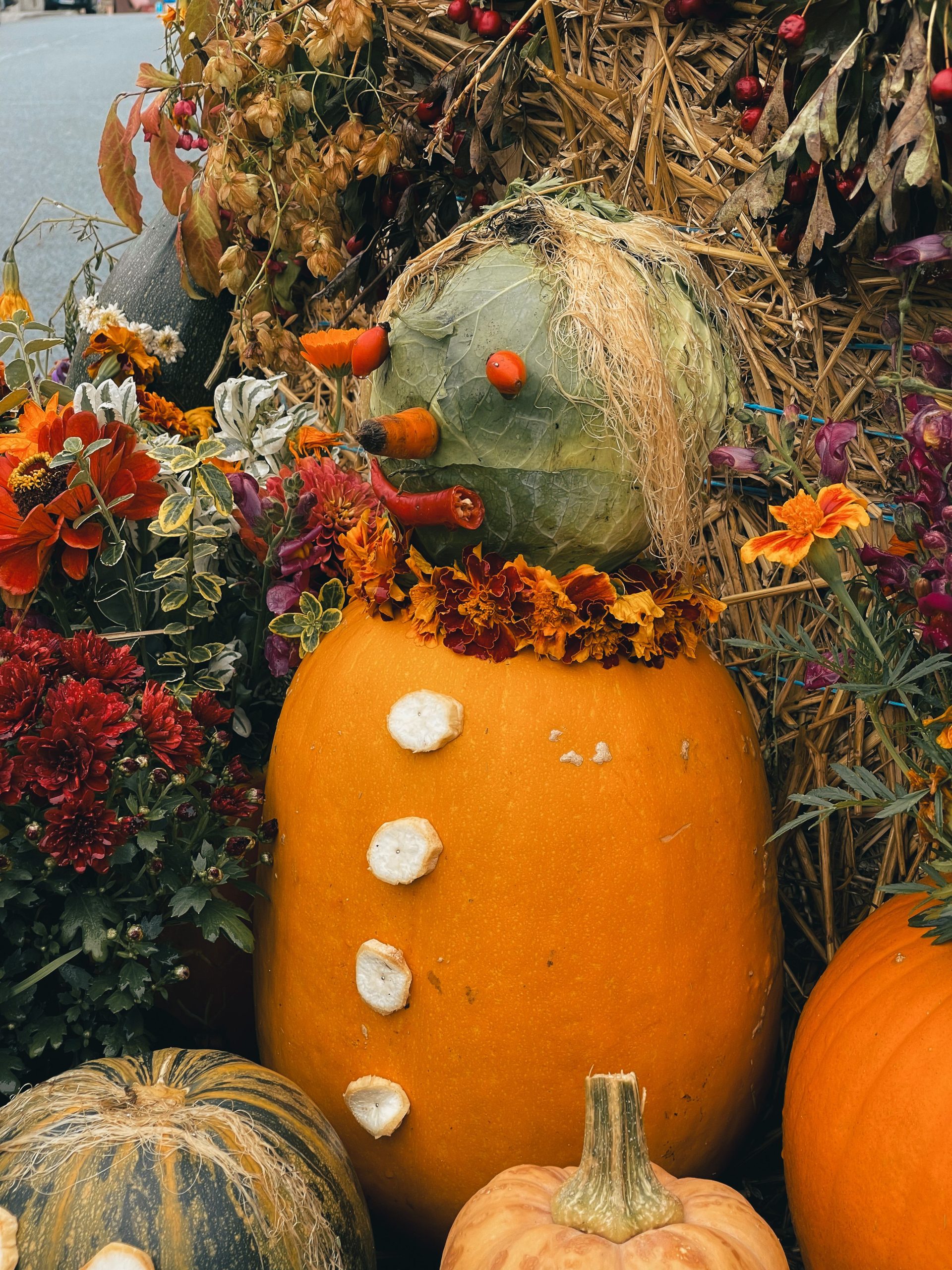 Festive autumn display featuring a whimsical pumpkin scarecrow adorned with flowers and vegetables.