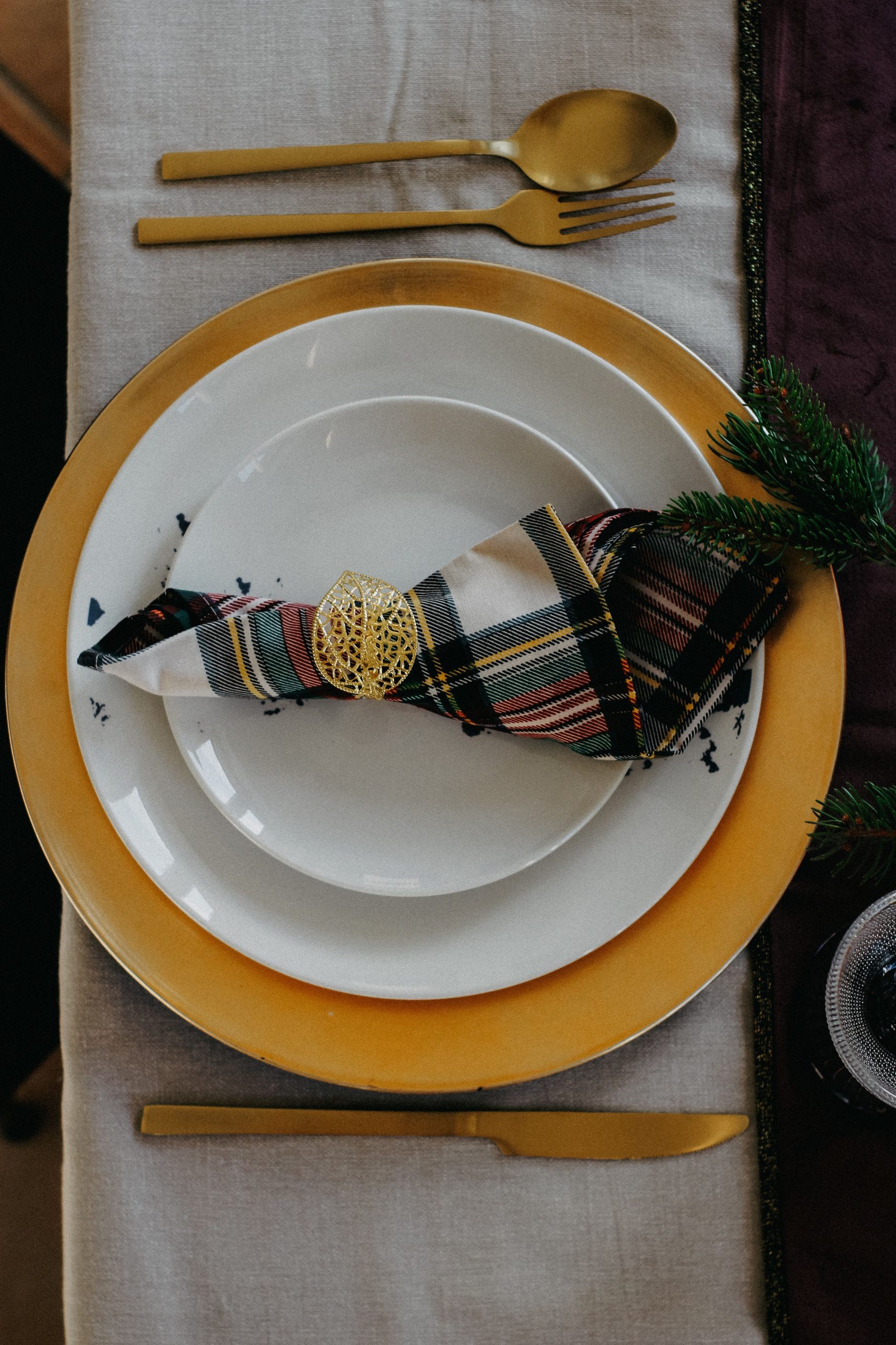Festive table setting featuring gold accents, plaid napkin, and spruce branch detail.