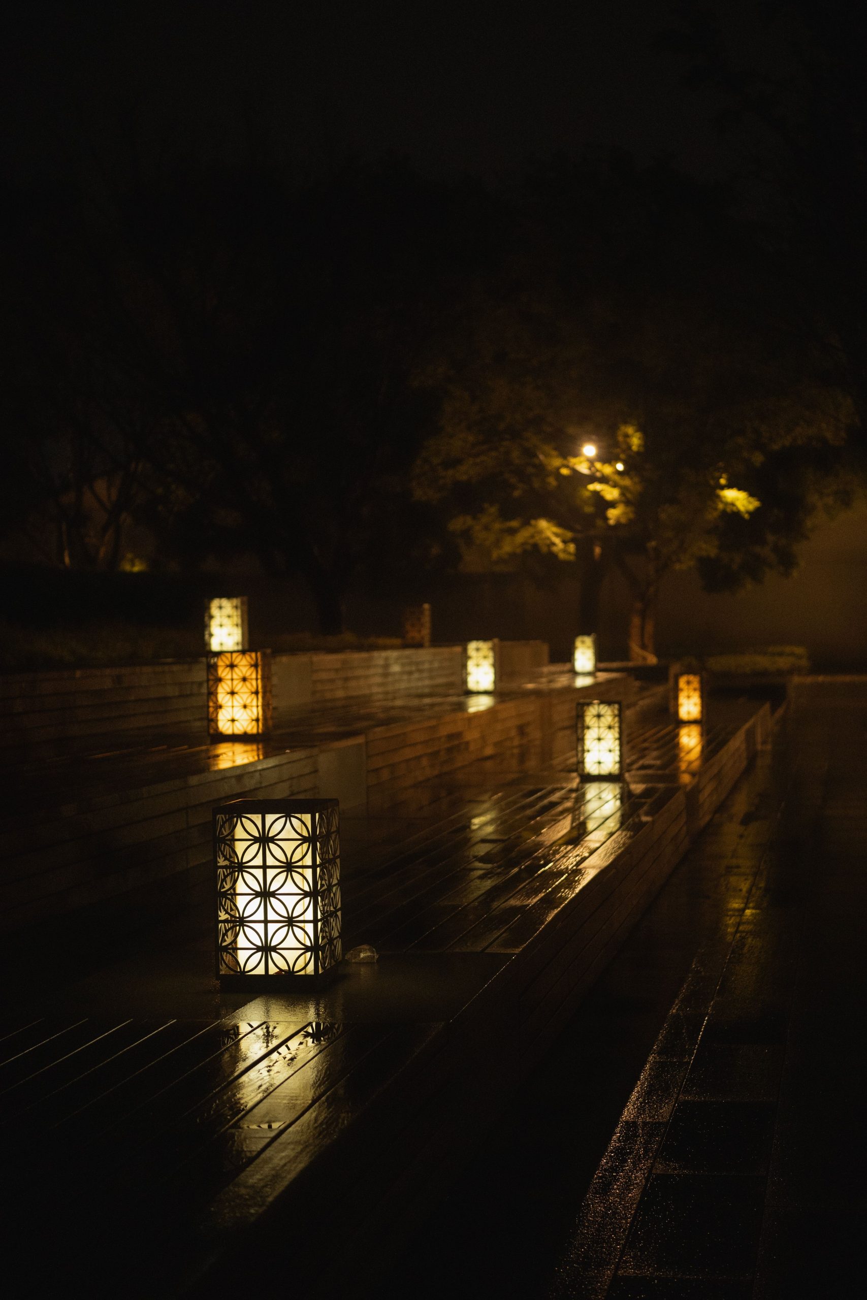 Serene nighttime scene of glowing lanterns lining a wooden path, reflecting on wet surfaces.