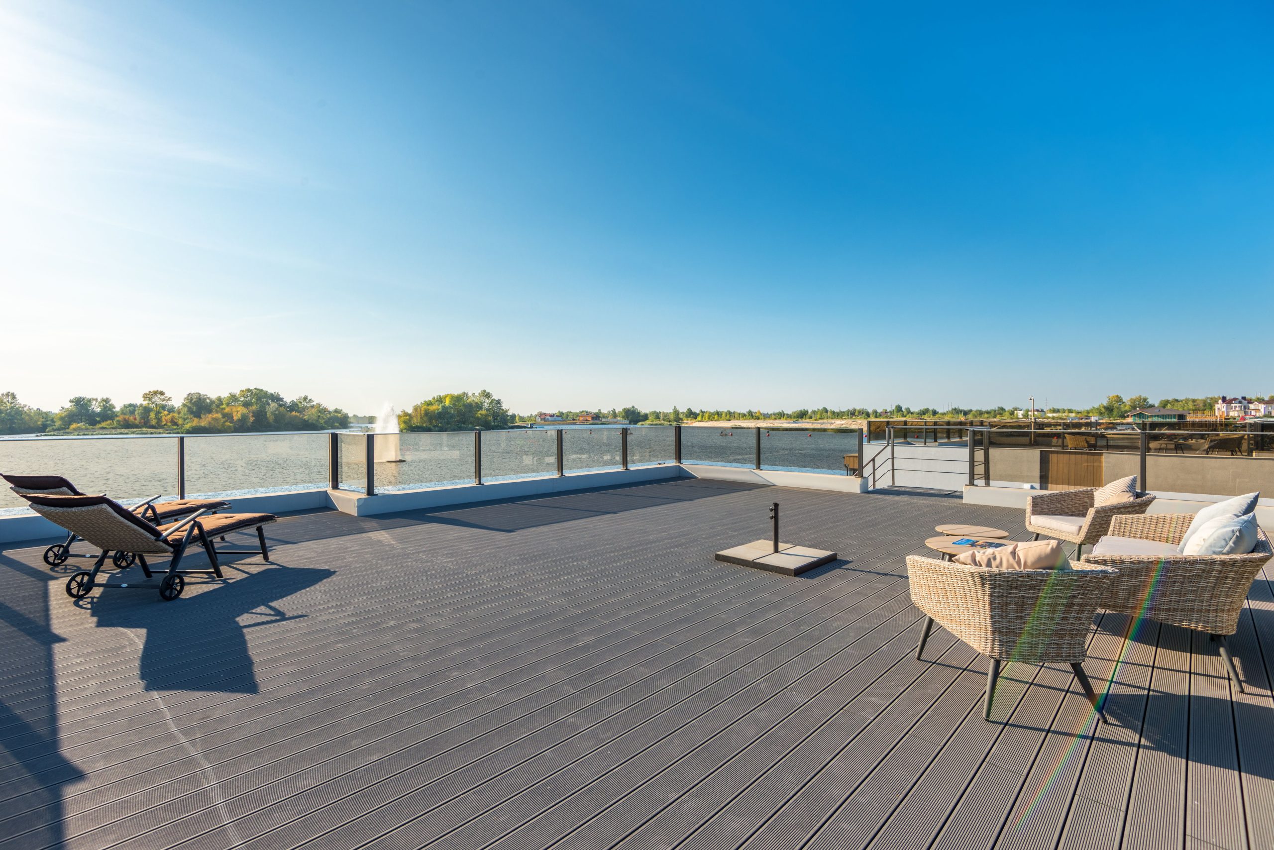 Spacious, sunny waterfront deck with modern wicker seating and lounge chairs overlooking a serene lake.