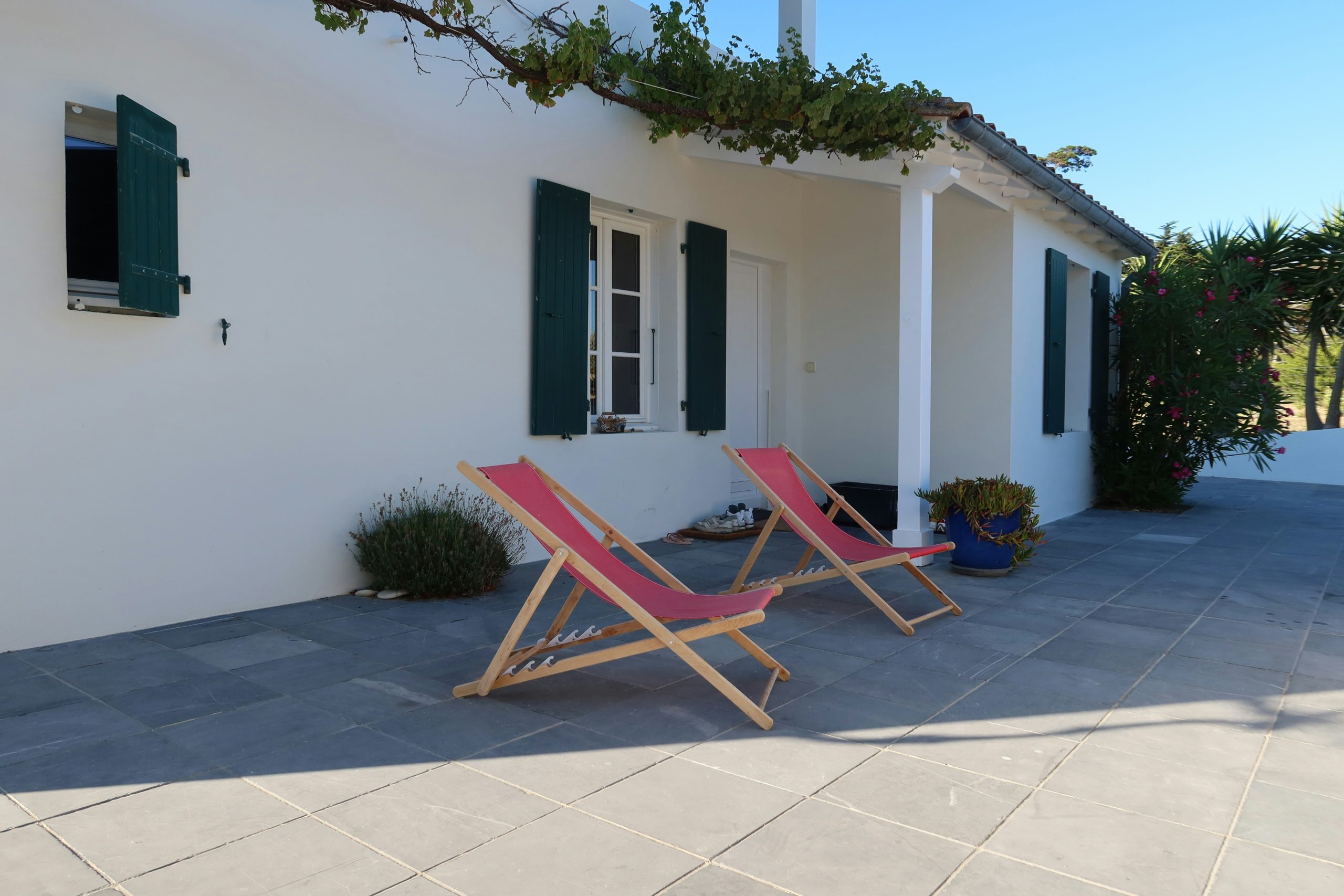 Two red canvas deck chairs sit on a gray tiled patio in front of a crisp white house with dark green shutters, shaded by a leafy vine canopy and flanked by potted plants for a relaxed summer terrace s