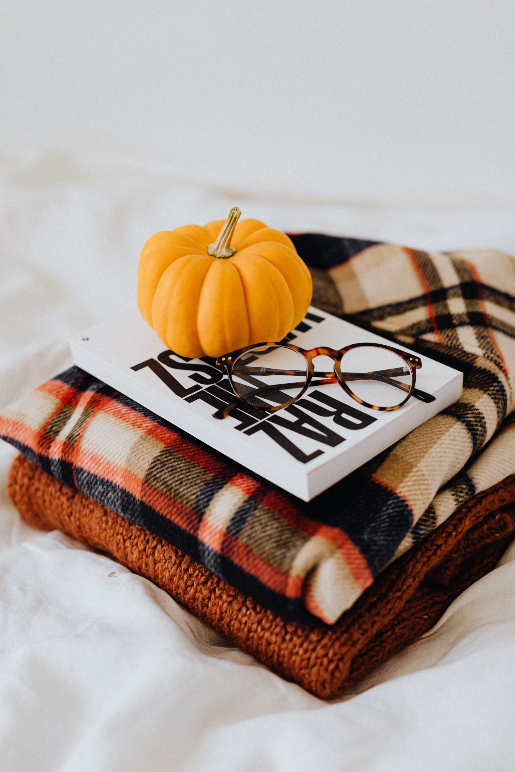 Warm autumn setting featuring a small pumpkin, eyeglasses, and a plaid fabric stack.