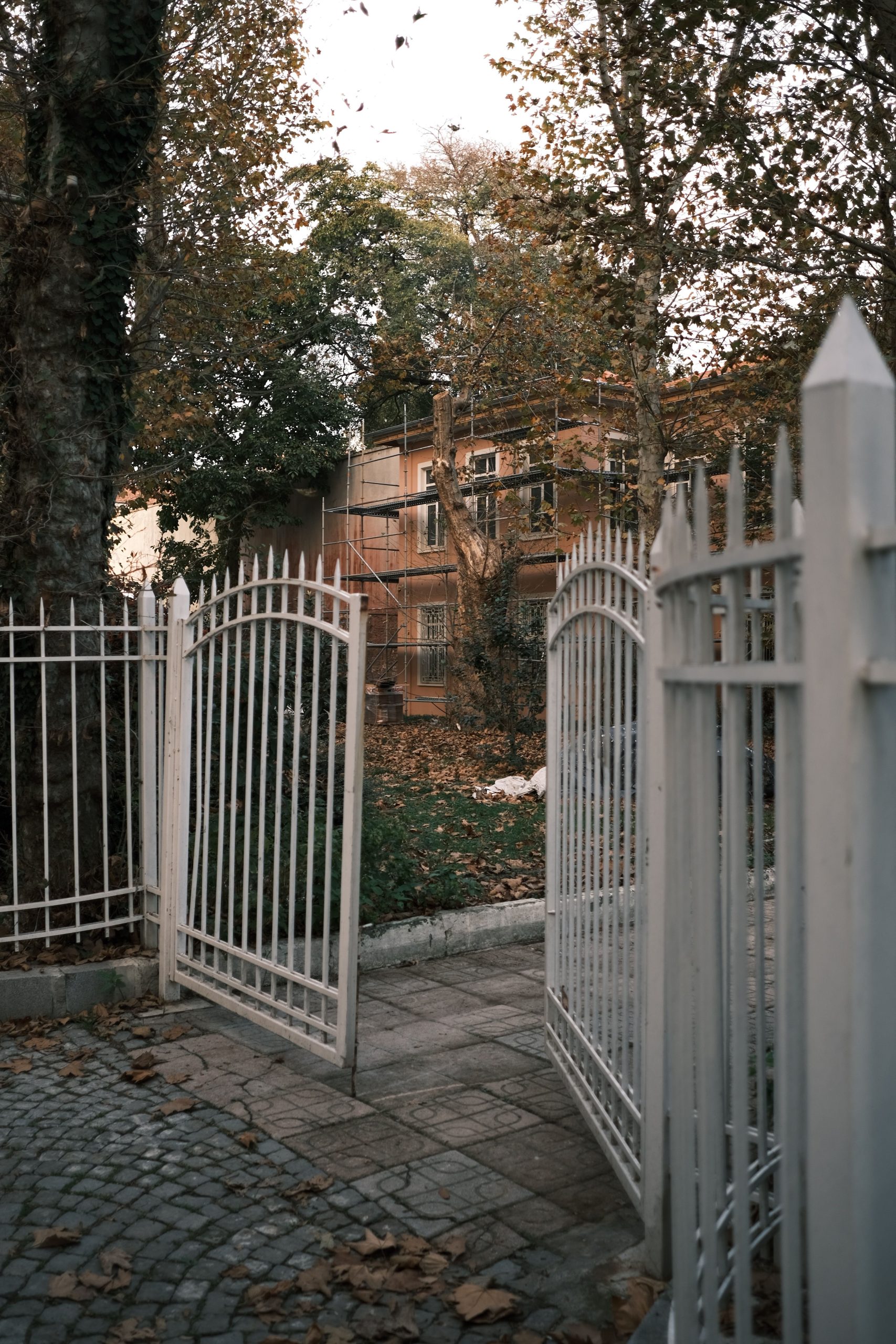 White metal gates open to a house surrounded by autumn trees and fallen leaves.