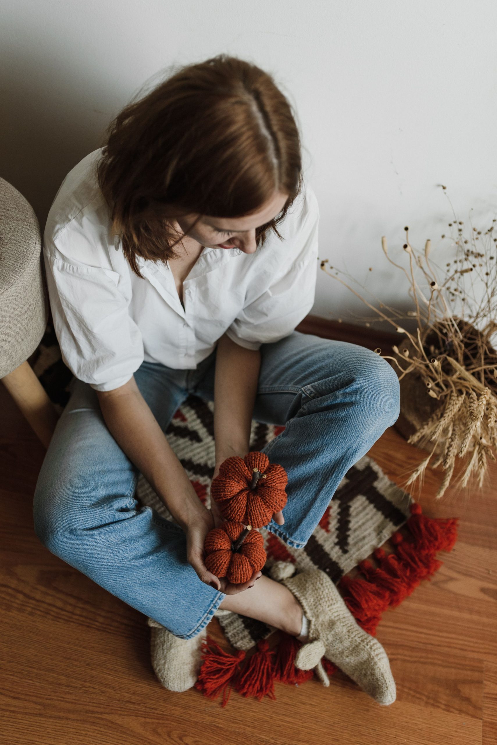 Woman sitting cross-legged with knitted pumpkins on wooden floor, embracing fall vibes.