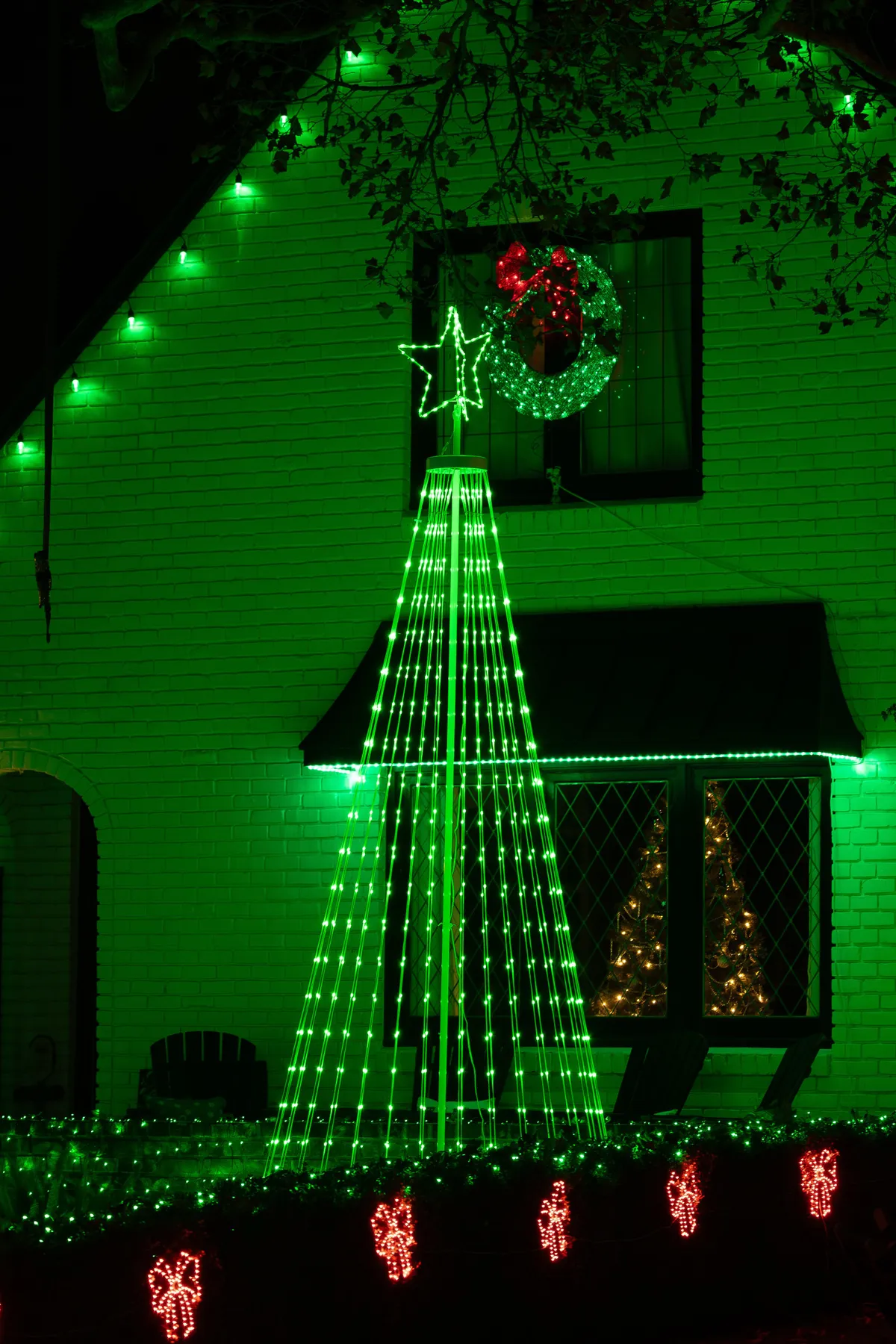 Bright green and red Christmas lights adorn a house at night, featuring a lit tree and wreath.
