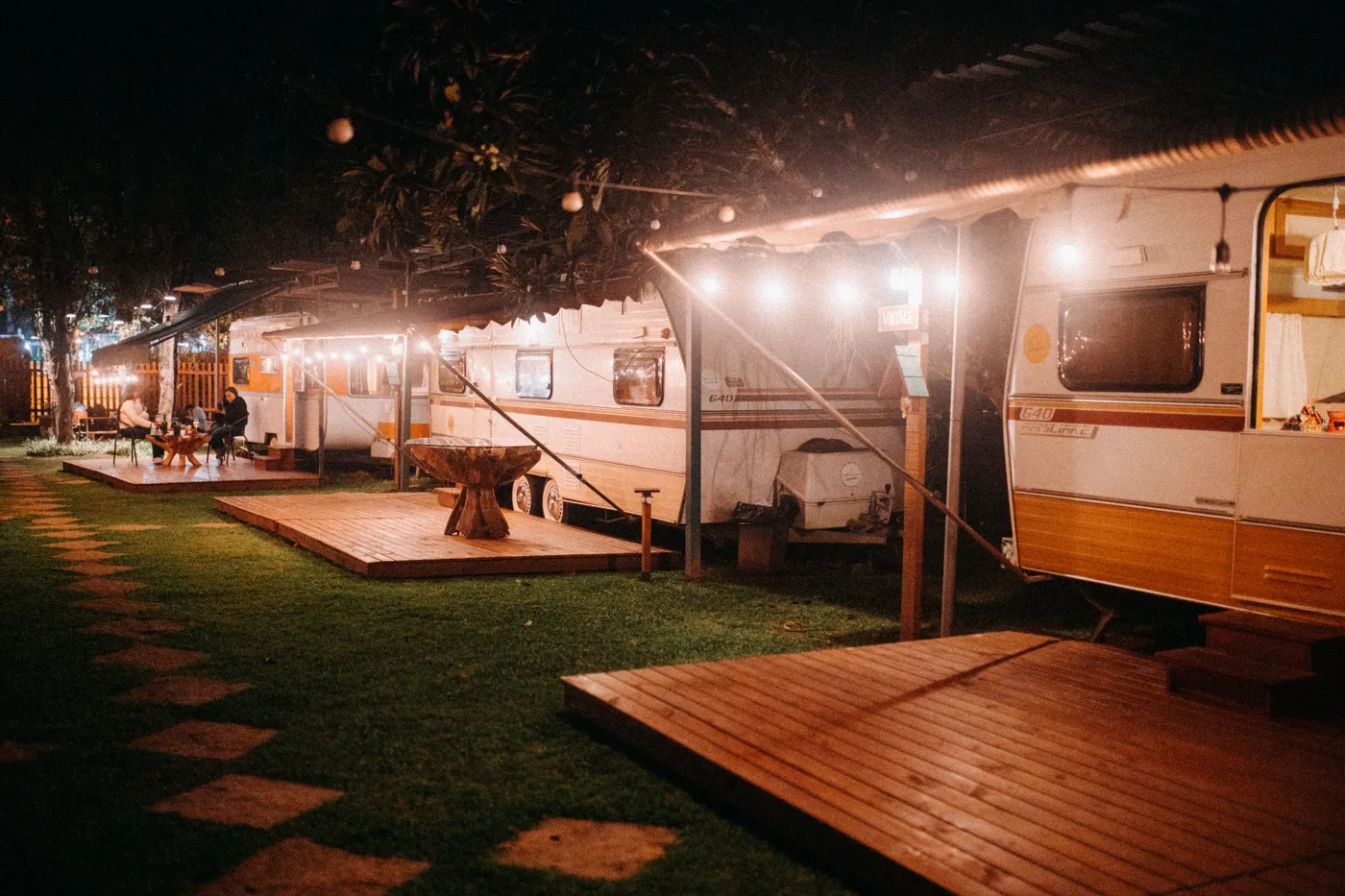 Campground with row of trailers and anonymous female travelers interacting at table under canopy and shiny lights at dusk