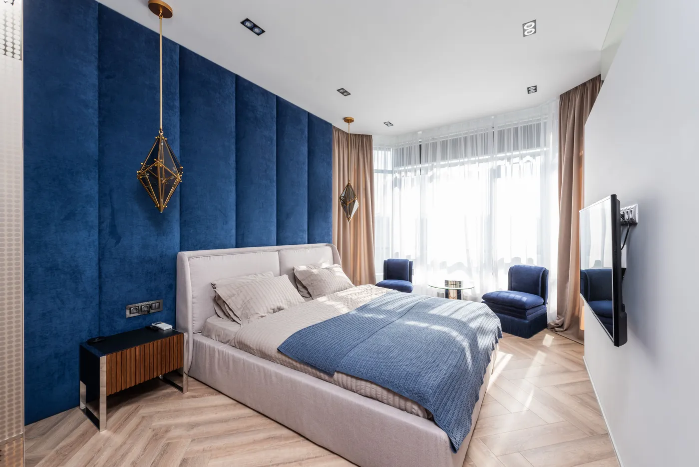 Comfortable bed with cushions and blanket placed near blue wall against TV in spacious bedroom with armchairs and table near window
