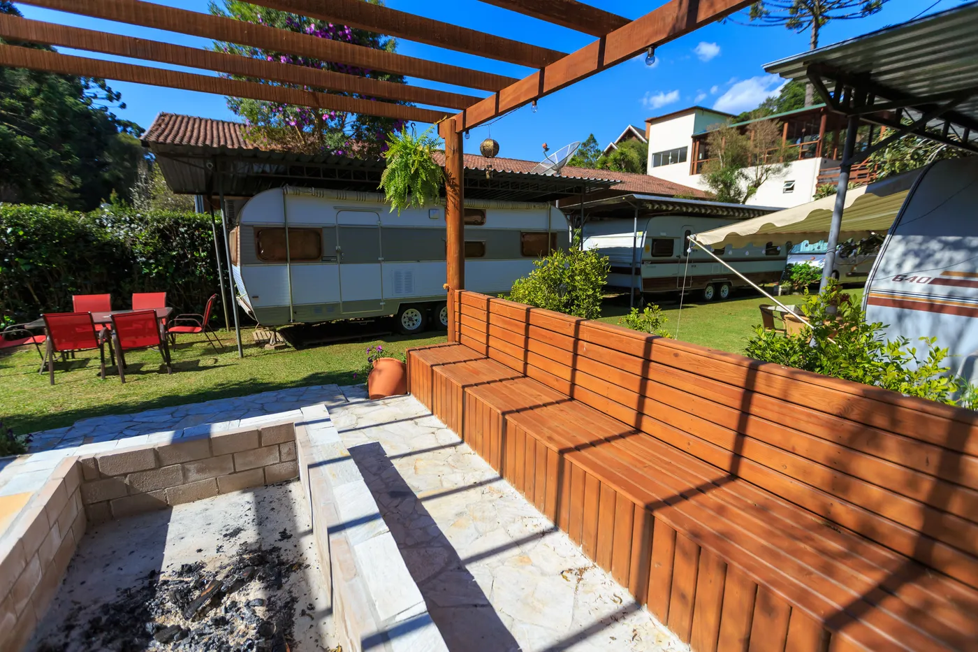 Cozy campground featuring wooden benches, caravans, and a fire pit under a sunny sky.