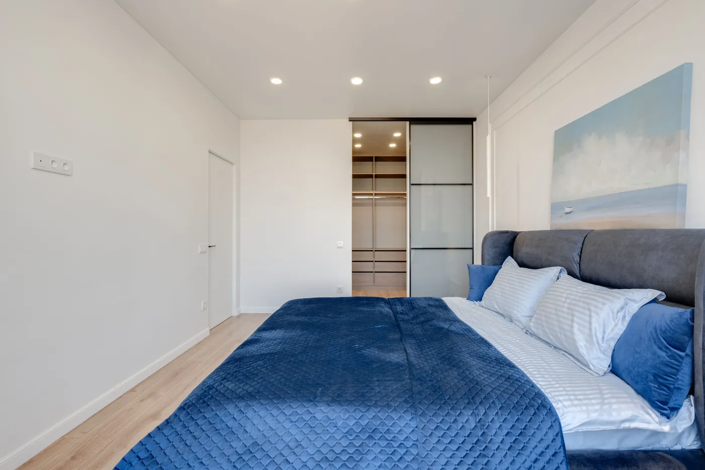 Spacious bedroom featuring a modern design with blue bedding, an abstract painting, and a sliding door cabinet.