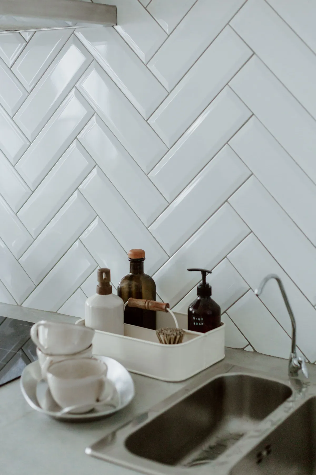 Stylish kitchen sink area with cleaning products and cups on a tiled counter.